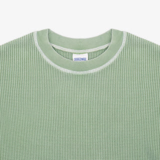 Close-up of a light sage green waffle-knit crewneck sweater with a white ribbed collar trim and a visible neck label reading 'SWEATWEAR'.