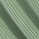 A close-up of a light sage green knit fabric with a textured, ribbed pattern and soft diagonal folds.