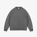 A dark gray crewneck sweatshirt with raglan sleeves, laid flat against a light gray background. A small circular logo patch is visible on the left sleeve cuff.