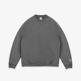 A dark gray crewneck sweatshirt with raglan sleeves, laid flat against a light gray background. A small circular logo patch is visible on the left sleeve cuff.