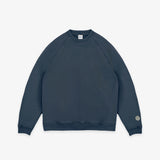 A dark slate blue crewneck sweatshirt with raglan sleeves, shown flat against a light gray background. A small circular logo is printed on the left sleeve cuff.