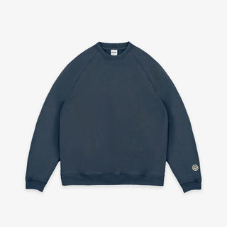 A dark slate blue crewneck sweatshirt with raglan sleeves, shown flat against a light gray background. A small circular logo is printed on the left sleeve cuff.