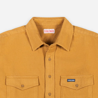 Close-up of a mustard yellow button-up shirt with a pointed collar, two chest pockets, and a visible Dubbleware neck label and a small chest patch logo.