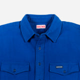 A close-up of a royal blue button-down shirt by Doubleware, featuring a classic collar, dark buttons, and two buttoned chest pockets on a light gray background.