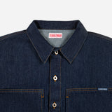 Close-up of a dark indigo denim button-up shirt by Dubble Wade, showing the collar, front buttons, chest pockets, and a small brand tag on the right chest pocket.
