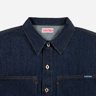 Close-up of a dark indigo denim button-up shirt by Dubble Wade, showing the collar, front buttons, chest pockets, and a small brand tag on the right chest pocket.
