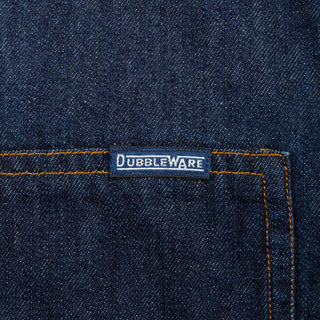 Close-up of dark indigo denim fabric with orange contrast stitching and a small rectangular Dubbleware label near the pocket.