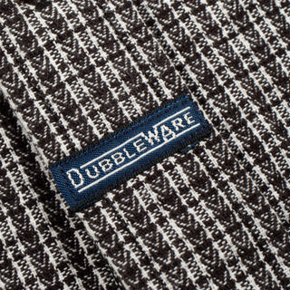 Close-up of a woven knit fabric with a dark brown and off-white textured pattern, featuring a blue embroidered label with the brand name 'OUBLEWARE' in white.