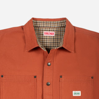 Close-up of a Doubleware work shirt in rust orange, showing the collar with a brown plaid lining, snap-button front, and a small white brand patch on the chest pocket.