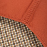 Close-up detail of a garment corner showing a rust-orange woven fabric body meeting a brown, beige, and black plaid lining.