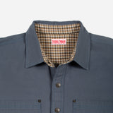 Close-up of a Doubleware button-up shirt in slate blue, showing the inner plaid lining of the collar and the brand label.