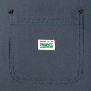 Close-up of a blue canvas work shirt pocket with two metal snap buttons and a white Dubbleware brand label stitched in the center.