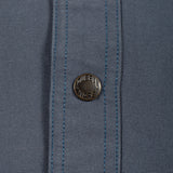 Close-up of a slate blue woven fabric with a single round metal button featuring the 'MURDER WARE' brand name.
