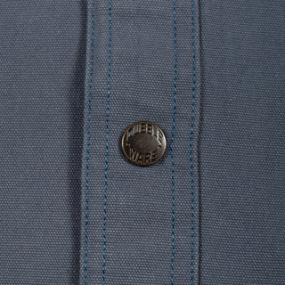 Close-up of a slate blue woven fabric with a single round metal button featuring the 'MURDER WARE' brand name.